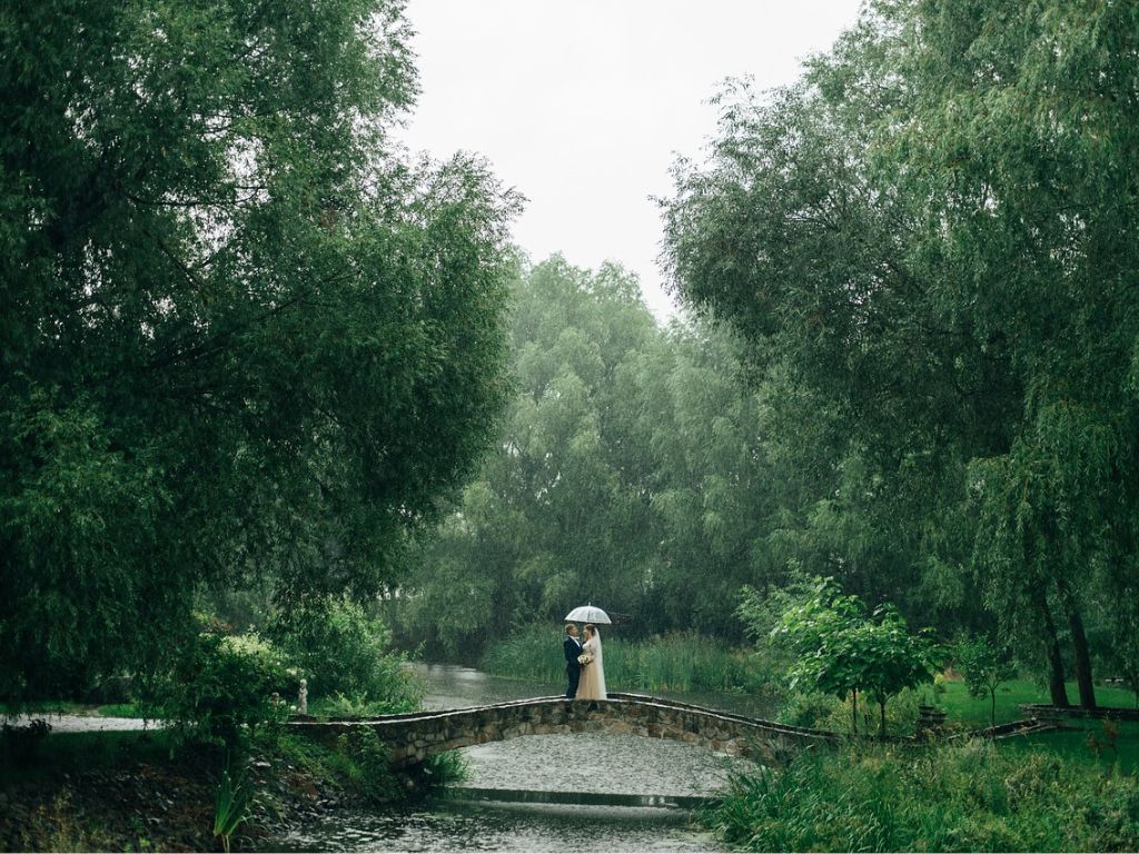 Auch bei Regen lassen sich romantische Hochzeitsbilder erzielen, wie das Brautpaar unterm Regenschirm im Park.